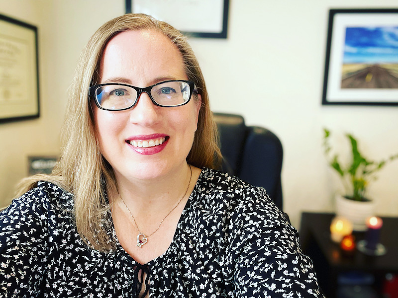 elizabeth potts weinstein, a smiling woman 
          with glasses, in front of out-of-focus background of plant, candles, and wall art